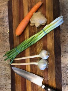 chef's knife, 2 garlic heads, 1 bunch of green onions, 1 piece of ginger, and 1 large carrot on a cutting board