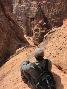 man crouching and looking down at the petra treasury, a building carved into a cliff face